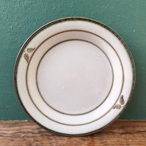 Puede incluir: Un plato de cerámica blanco con un borde oscuro y dos círculos concéntricos marrones. El plato tiene un diseño sencillo con dos conjuntos de patrones en forma de hoja. El plato está sobre un fondo verde y una superficie de madera.