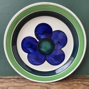 Könnte beinhalten: Ein runder Keramikteller mit einem weißen Mittelteil mit einem blauen Blumenmuster. Die Blume hat fünf Blütenblätter und ein grünes Zentrum. Der Teller hat einen grünen und schwarzen Rand und einen grünen Rand.