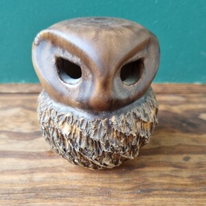 以下が含まれることがあります： 木製の小さな茶色のフクロウの置物。彫刻された質感のあるベースがあり、大きな空洞の眼窩と滑らかで丸みを帯びた頭部が特徴です。この置物は、おそらく室内装飾用の装飾品です。