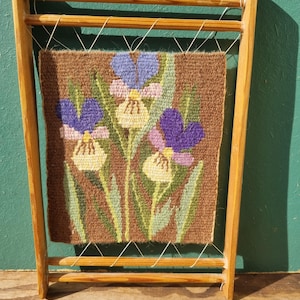 May include: A framed tapestry featuring a floral design of purple, blue, and yellow irises on a brown background. The tapestry is held within a light-colored wooden frame, showcasing a vintage craft aesthetic.