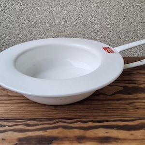 May include: A white enamel bedpan with a handle. The bedpan is round with a wide rim and a curved handle. The bedpan is sitting on a wooden surface.