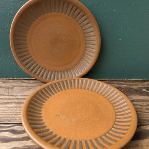 May include: Two brown ceramic plates with a textured, ridged design around the edges. The plates are circular and have a matte finish. The plates are displayed on a wooden surface.