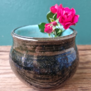 May include: A small, dark brown ceramic vase with a light blue glaze inside. The vase has a rounded shape and a slightly flared rim. A single stem of pink roses is placed inside the vase.