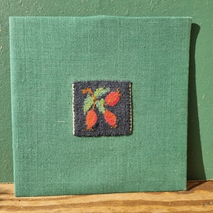 May include: A square, green fabric-covered item with a small, embroidered square in the center. The embroidered square features a dark background with red berries and green leaves. The item is displayed on a wooden surface.