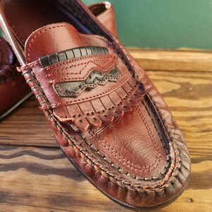 May include: A pair of brown leather loafers with a fringe detail and a black leather accent. The loafers have a braided trim around the edge of the sole.