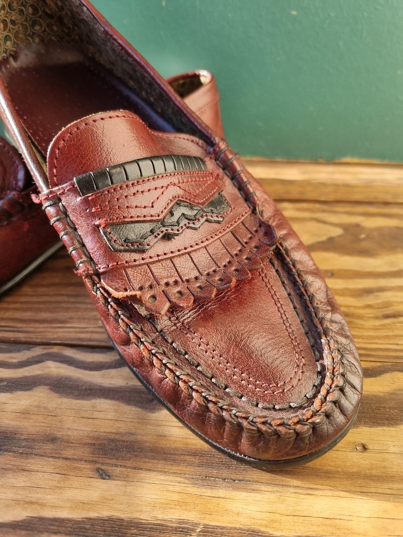 May include: A pair of brown leather loafers with a fringe detail and a black leather accent. The loafers have a braided trim around the edge of the sole.