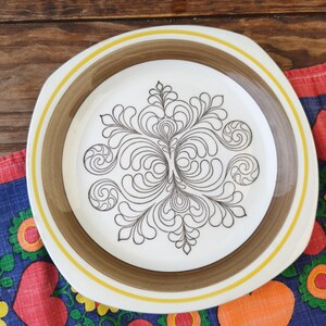 Puede incluir: Un plato cuadrado blanco vintage con un borde marrón y amarillo. El plato presenta un diseño floral negro y arremolinado en el centro. El plato está sobre una tela estampada colorida.