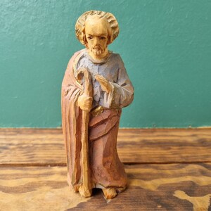 May include: A wooden figurine of a man with a halo, holding a staff. He is wearing a long robe with a blue and brown pattern. The figurine is carved in a realistic style and has a warm, natural finish.