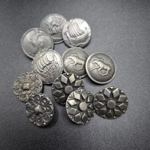 Könnte beinhalten: Eine Sammlung antiker, silberfarbener Knöpfe. Die Knöpfe weisen verschiedene Designs auf, darunter ein Wikingerschiff, ein Pferd sowie florale und geometrische Muster. Die Knöpfe sind rund und haben ein leicht gealtertes Aussehen.