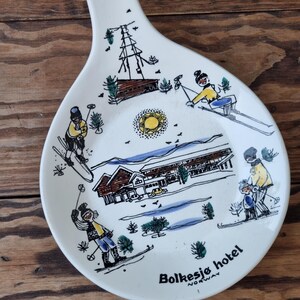 以下が含まれることがあります： ハンドルが付いた白いセラミックプレートで、スキーヤーとホテルの冬のシーンが描かれています。プレートは青と緑のデザインで、黄色の太陽と「Bolkesjø hotel NORWAY」の文字が書かれています。