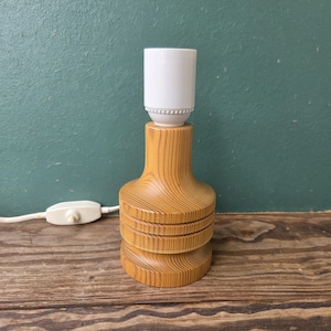 May include: A table lamp base crafted from light-coloured wood with a prominent grain pattern. The base features a cylindrical, tiered design with a white plastic light bulb socket at the top and a white electrical cord with an inline switch.