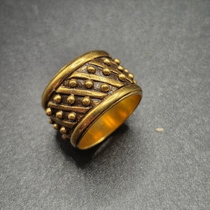 May include: A wide, gold-colored ring with a textured design. The ring features a series of horizontal and diagonal lines, interspersed with small, raised circular details. The interior of the ring is a smooth, polished gold. The ring is likely made of metal.