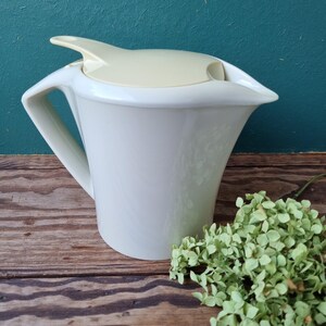 Peut inclure: Une cruche en céramique blanche avec un couvercle et une poignée crème. La cruche a un design incurvé et un bec verseur. L'arrière-plan est un mur turquoise et une surface en bois. Un petit bouquet de fleurs vertes séchées est à droite.