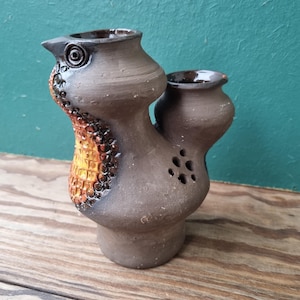 May include: A brown ceramic bird-shaped vase with two openings. The vase has a textured surface with yellow and orange accents. The vase has a small hole on the side.