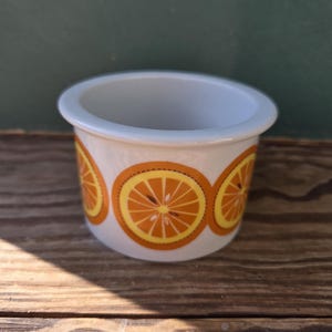 Può includere: Un piccolo contenitore in ceramica bianca con un bordo azzurro. Il contenitore è decorato con disegni di fette d'arancia arancioni e gialle. Il contenitore è su una superficie di legno.