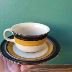Può includere: Un set da tè in ceramica a strisce marroni e bianche. La tazza ha un bordo largo e un manico. Il piattino ha un bordo marrone e un centro bianco.