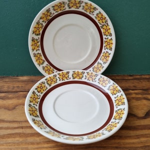 May include: Two white ceramic saucers with a brown rim and a yellow and brown floral pattern. The saucers have a center well for a cup.