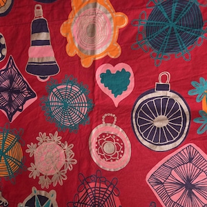 May include: A red fabric with a repeating pattern of colorful, stylized Christmas ornaments. The ornaments include bells, baubles, and snowflakes in shades of blue, pink, orange, and teal. The design is hand-drawn and whimsical.