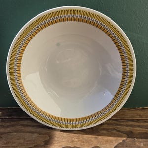 Puede incluir: Un cuenco redondo de cerámica blanquecina con un borde decorativo. El borde presenta un patrón repetitivo de elementos amarillos y azules. El cuenco está sobre un fondo verde oscuro y una superficie de madera.