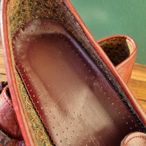 May include: Close-up of the inside of a burgundy leather shoe. The insole is burgundy leather with a pattern of small holes. The lining is a patterned fabric with a burgundy, green, and gold design.