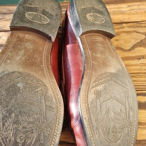 May include: Close-up of the soles of a pair of burgundy leather loafers. The soles are brown leather with a detailed design of a tree and a building. The text "MADE IN ITALY" is visible on the right shoe.