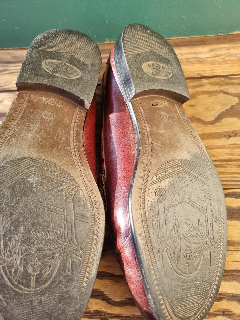 May include: Close-up of the soles of a pair of burgundy leather loafers. The soles are brown leather with a detailed design of a tree and a building. The text "MADE IN ITALY" is visible on the right shoe.