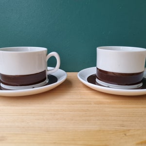 Può includere: Due tazze da caffè in ceramica bianca con bande marroni intorno alla parte superiore. Le tazze sono su piattini bianchi.