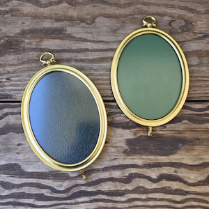 May include: Two oval-shaped picture frames with gold-colored metal rims. One frame has a dark green interior, and the other has a black interior. Each frame has a small loop at the top and a hook at the bottom.