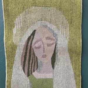 May include: Woven wall hanging featuring a stylized portrait of a person with closed eyes. The artwork uses muted tones of green, pink, and gray. The piece is rectangular and is held by two wooden clothespins.