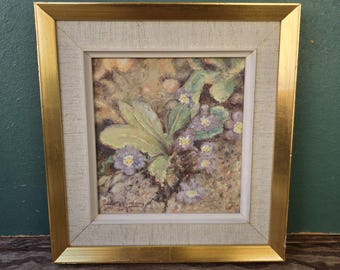 May include: A framed painting featuring a cluster of light purple flowers with yellow centers and green leaves. The artwork is encased in a gold-toned frame with a textured, off-white fabric matting surrounding the central image.