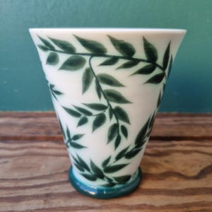 May include: A white ceramic vase with a dark green leaf pattern. The vase has a flared top and a teal-colored base. The leaves are painted in a detailed, naturalistic style, creating a decorative floral design.