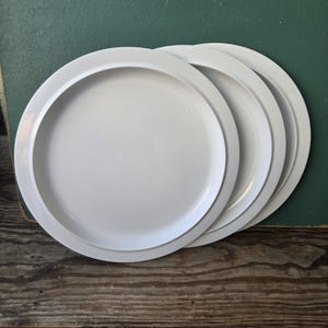 May include: Three plain white circular plates with a smooth finish and a wide, flat rim. The plates are stacked slightly offset from one another, revealing the consistent round shape and clean, solid white color of each individual dinner plate.