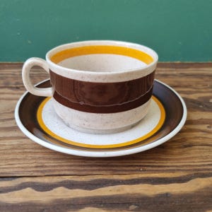 May include: A brown and white teacup and saucer with a yellow stripe. The cup and saucer have a speckled glaze.