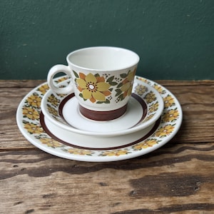 May include: A ceramic tea cup, saucer, and plate set featuring a cream-colored base with a repeating pattern of yellow and orange flowers, green leaves, and a solid dark brown band around the base of the cup and the center of the plates.