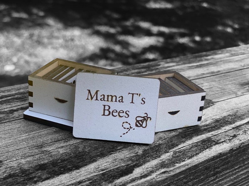 May include: A wooden box with a white lid that says "Mama T's Bees" with a bee illustration. The box is open and the lid is propped up.