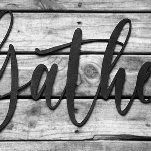 May include: Black metal word art spelling out the word "gather" in a cursive font. The word art is mounted on a wood background.