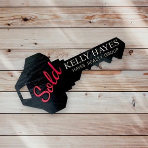 May include: A black wooden key-shaped sign with the word "Sold" in red cursive lettering and the text "Kelly Hayes Hayes Realty Group" in white lettering.