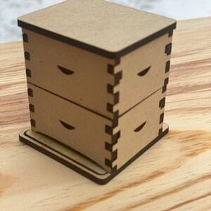 May include: A wooden beehive model with three stacked boxes and a base. The boxes have a dark brown trim and a cutout design on the front.