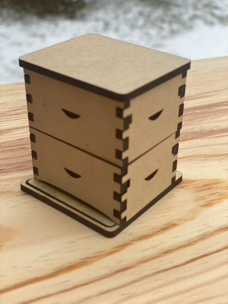 May include: A wooden beehive model with three stacked boxes and a base. The boxes have a dark brown trim and a cutout design on the front.