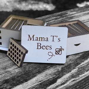 May include: A wooden beehive with a sign that reads "Mama T's Bees" with a bee and honeycomb design. The beehive is made of wood and has a natural finish.