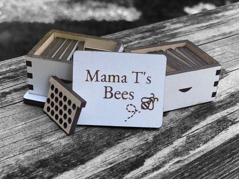 May include: A wooden beehive with a sign that reads "Mama T's Bees" with a bee and honeycomb design. The beehive is made of wood and has a natural finish.