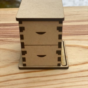 May include: A small wooden box with two drawers. The box is made of light brown wood and has a dark brown trim. The drawers have a simple design with a small cutout on the front.