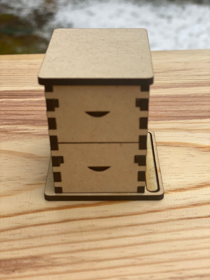 May include: A small wooden box with two drawers. The box is made of light brown wood and has a dark brown trim. The drawers have a simple design with a small cutout on the front.
