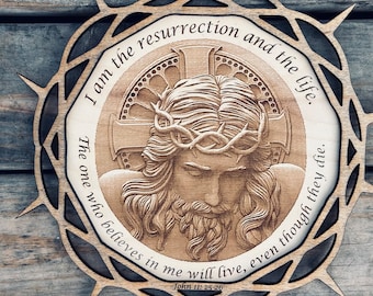 May include: Wooden religious wall art featuring a detailed relief of Jesus wearing a crown of thorns, surrounded by a circular frame with text from John 11:25-26. The artwork is in shades of brown and tan, with a decorative, spiky outer border.