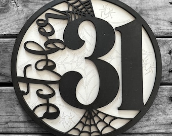 Op de afbeelding: Een rond zwart-wit Halloween-bord met de nummers 31 en een spinnenwebontwerp. Het bord zegt "Boo" in een decoratief lettertype.