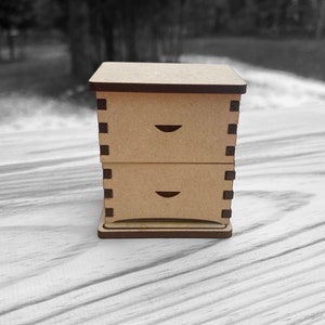 May include: A small wooden box with two drawers. The box is made of light brown wood and has a dark brown trim. The drawers have a simple design with a small cutout on the front.