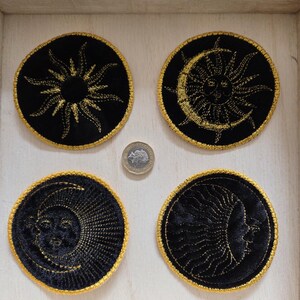 May include: Four circular black fabric patches with gold embroidered celestial designs, including suns and moons with faces. A small silver coin is placed in the centre for scale, showing the relative size of the embroidered circular patches.