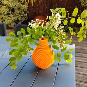 Peut inclure: Un vase orange vif avec un motif en spirale contient un bouquet de fleurs blanches avec des feuilles vertes. Le vase est posé sur une table grise avec une terrasse en bois en arrière-plan.