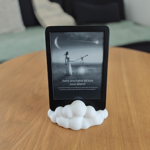 Op de afbeelding: Een zwarte e-reader met een scherm waarop een persoon door een telescoop naar de maan en een vallende ster kijkt. De tekst luidt "Votre prochaine lecture vous attend". Het apparaat staat in een witte, wolk-vormige standaard.