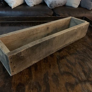 Puede incluir: Una jardinera rectangular de madera con un acabado desgastado gris-marrón. La caja está construida con tablas de madera y tiene cabezas de clavos visibles. El interior es abierto y la caja se encuentra sobre una superficie de madera oscura.